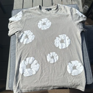 Beige t-shirt med vita tryck Denim Tears - Snygg beige t-shirt från Denim Tears i bomull med coola vita tryck i form av cirklar över hela plagget, både fram och bak samt på ärmarna. Klassisk rund hals och korta ärmar. Perfekt för dig som gillar streetwear och unik design.