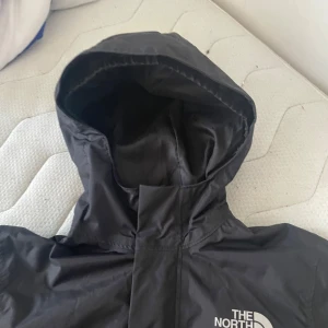 Svart vindjacka från The North Face - Svart vindjacka från The North Face med huva och elastiska muddar vid ärmsluten. Jackan har en clean look med logga på bröstet och är tillverkad i lätt polyester som står emot vind och regn. Perfekt för dig som vill ha en stilren jacka med funktion.
