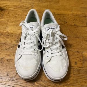 Vita Adidas sneakers med svarta ränder - Snygga vita Adidas sneakers med klassiska svarta ränder på sidorna. Skorna har rund tå, platt sula och snörning framtill. Tillverkade i canvasmaterial med vit yttersula och diskret Adidas-logga på plösen. Perfekt för dig som gillar stilrena och sportiga skor.