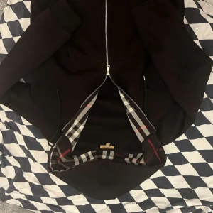 Svart Burberry bomberjacka XL, sitter som en L, krympt lite - Säljer en svart bomberjacka från Burberry i storlek XL. Jackan har klassiskt rutigt foder, silverfärgad dragkedja och ribbade muddar. Materialet är mjukt och känns lyxigt, perfekt för dig som vill ha en stilren jacka med ikonisk Burberry-touch.