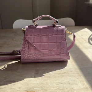Ljuslila croco handväska med axelrem - Snygg ljuslila handväska med croco-mönster och guldfärgade detaljer. Väskan har både handtag och justerbar axelrem, samt ett lock med magnetknäppning. Materialet är glansigt syntet och insidan är svart. Perfekt för dig som gillar trendiga accessoarer.