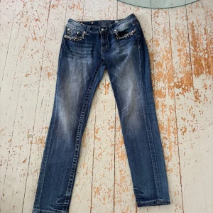 Miss Me blå jeans med pärldetaljer - Snygga blå jeans från Miss Me med pärl- och nitedetaljer på både fram- och bakfickor. Jeansen har en klassisk femficksdesign, låg midja och raka ben. Materialet är slitstarkt denim med coola slitningar och tvättade effekter för en trendig look.