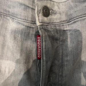 Grå Dsquared2 jeans med slitningar - Snygga grå jeans från Dsquared2 med slitna detaljer och färgstänk för en cool, avslappnad vibe. Klassisk femficksmodell med Dsquared2-logga på röd etikett och patch bak i midjan. Raka ben och normal passform, perfekt till sneakers.