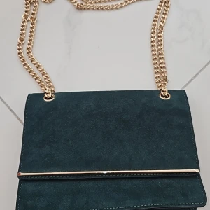 Mörkgrön mocka axelväska New Look - Snygg mörkgrön axelväska från New Look i mjuk mocka med guldfärgad kedja och metall-detalj på locket. Perfekt storlek för mobilen och det viktigaste. Magnetlås och ett fack på insidan. Stilren och trendig design som lyfter din outfit.