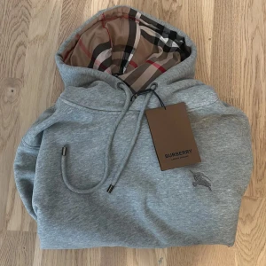 Grå Burberry hoodie  - Snygg grå hoodie från Burberry i storlek M med klassiskt rutigt foder i huvan och broderad logga på bröstet. Tröjan har snörning vid huvan, ribbade muddar och en stor magficka. Perfekt för dig som gillar stilrena designer med lyxig touch.