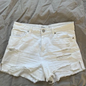 Vita jeansshorts från FB Sister - Snygga vita jeansshorts från FB Sister i storlek L. Modellen är högmidjad med råa, fransiga kanter och slitna detaljer framtill för en cool vibe. Klassisk femficksdesign och dragkedja med knapp. Perfekta för sommarens alla äventyr! I mycket fint skick!