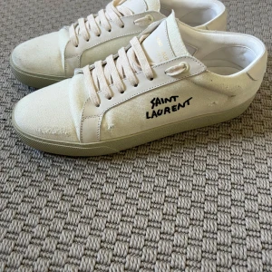 Saint Laurent vita sneakers med logga - Snygga vita sneakers från Saint Laurent med handskriven logga i svart på sidan. Skorna har rund tå, platt sula och snörning. Tillverkade i canvas och skinn med coola slitna detaljer för en avslappnad vibe. Perfekta för dig som gillar stilren design med edge.