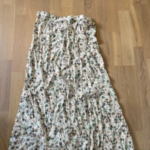 Blommig långkjol  - Långkjol från H&M i storlek 40 med blommigt mönster i beige, grönt och svart. Kjolen har en ljus bas och stängs med dragkedja i sidan. 