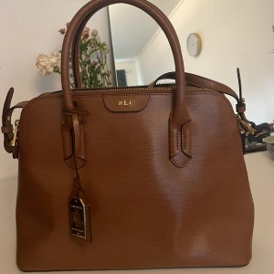 Brun handväska från Ralph Lauren - Snygg brun handväska från Ralph Lauren med stilrena detaljer i guld och präglad struktur. Väskan har två rejäla handtag, dragkedja upptill och en tag med RL-logga. Perfekt storlek för allt du behöver ha med dig och passar till många olika looks.