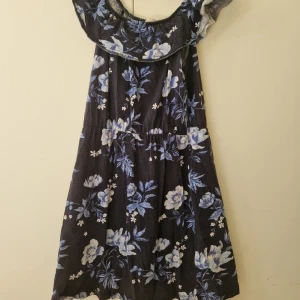 Blommig offshoulder klänning från H&M - Klänning från H&M med offshoulder och volang upptill. Mörkblå botten med stora blommor i blått och vitt. Perfekt för sommaren