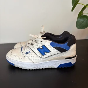New Balance 550 sneakers vit/blå - Säljer ett par New Balance 550 sneakers i vitt och blått med svarta detaljer. Skorna har snörning, perforerade paneler och en platt sula med blå inslag. Ikonisk N-logga på sidan, perfekt för dig som gillar retrostil.