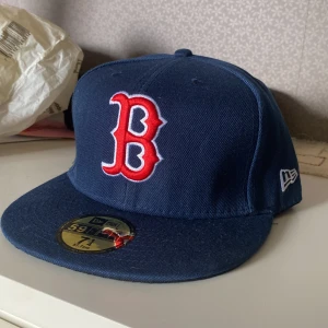 Blå Boston Red Sox keps New Era - Säljer en klassisk mörkblå Boston Red Sox 59FIFTY keps från New Era. Kepsen har en röd och vit broderad B-logga framtill, MLB-logga bak och New Era-logga på sidan. Platt skärm och stilren design, perfekt för dig som gillar baseball eller streetwear.