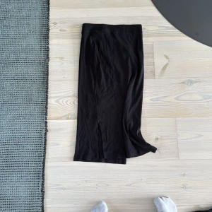 Svart långkjol från Name It - Svart ribbad långkjol från Name It i mjuk bomull. Kjolen har en liten slits framtill som ger en snygg detalj och elastisk midja för skön passform. Perfekt för dig som gillar en enkel men stilren look.