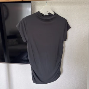 Grå kortärmad T-shirt från Cubus  - Mörkgrå T-shirt från Cubus med hög hals och korta ärmar. På båda sidorna finns de en rynkdetalj som är en perfekt detalj. T-shirten är tajt i passform och sitter väldigt fint på kroppen. Helt ny och har endast tagit av prislappen, säljer då den inte kommit till användning. 