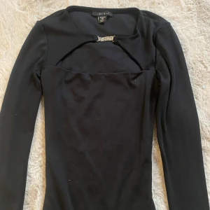 Svart långärmad topp med strassdetalj - Supersnygg svart långärmad topp från Amisu i storlek XS. Toppen har en cool cut-out vid halsen med en glittrande strassdetalj som ger extra edge. Mjuk och stretchig polyester som sitter slim på kroppen. Perfekt för dig som gillar att sticka ut med stil!