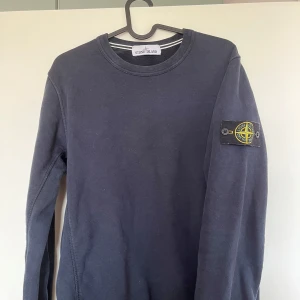 Mörkblå sweatshirt från Stone Island - Säljer en mörkblå sweatshirt från Stone Island med klassisk rund hals och lång ärm. Tröjan har den ikoniska Stone Island-patchen på vänster ärm och är tillverkad i mjuk bomull. Perfekt för dig som gillar streetwear och snygga detaljer.