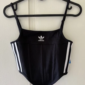 Svart korsett-topp från Adidas S - Supersnygg svart korsett-topp från Adidas med ribbad struktur och vita ränder längs sidorna. Toppens design har smala axelband och Adidas-logga framtill. Den är croppad och har en cool, formad nederkant som ger en edgy vibe.