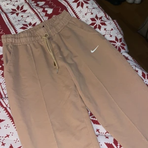Beige mjukisbyxor från Nike - Säljer ett par bekväma beige mjukisbyxor från Nike med elastisk midja och snörning. Byxorna har en avslappnad passform och är perfekta för en chill dag hemma. Den klassiska Nike-loggan syns på benet.