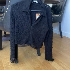 Spetskofta med dragkedja - Säljer en svart croppad kofta från Bershka med dragkedja framtill. Koftan är gjord i spetsmaterial som ger en cool och edgy look. Den har krage och långa ärmar, perfekt att styla över ett linne eller topp. Passar dig som gillar att sticka ut med detaljer.