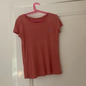Korallrosa t-shirt från Polo Ralph Lauren - Snygg korallrosa t-shirt från Polo Ralph Lauren med klassisk blå broderad logga på bröstet. Modellen är enkel med rund hals och korta ärmar, perfekt för chill dagar. Mjuk bomull som känns skön mot huden och har en normal passform.