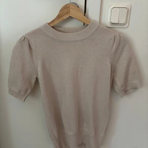 Beige stickad topp med puffärm - Snygg beige stickad topp med korta puffärmar och rund hals. Mjuk och skön kvalitet som ger en chill och stilren vibe. Perfekt att matcha med jeans eller kjol för en enkel men trendig look.