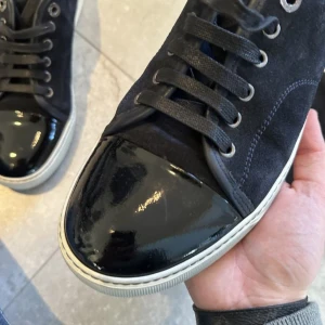 Svarta Lanvin sneakers med lackad tå - Snygga svarta sneakers från Lanvin med cool lackad tå och ovandel i mocka. Skorna har rund tå, snörning och vit platt sula som ger en clean vibe. Perfekta för dig som gillar stilrena detaljer och vill sticka ut med exklusiva material.