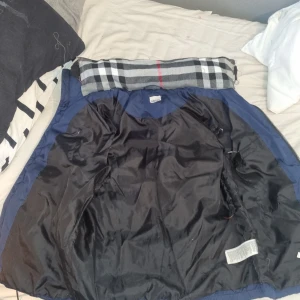 Burberry väst - Säljer en mörkblå dunväst med huva från Blueberry. Västen har två fickor med dragkedja och ett rutigt foder i svart, vitt och rött. Perfekt för kyliga dagar och har en snygg quiltad look. Passar dig som gillar streetwear och vill ha något varmt men ändå stilrent. Hemgjord burberry väst ifrån mig folket!