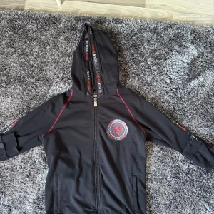Svart zip hoodie från Plein Sport - Svart hoodiejacka från Plein Sport i storlek XL med coolt tigertryck på bröstet och röda detaljer längs sömmarna. Jackan har hel dragkedja, huva med logotypband och två fickor framtill. Materialet är mjukt och skönt. Säljes då den inte använd längre, storlek XL men sitter som L, ny pris 6000 kr, pris kan diskuteras vid snabb affär👍