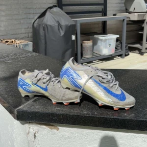 Nike Mercurial fotbollsskor silver/blå - Nike Mercurial fotbollsskor i silver med blå detaljer och stor blå Swoosh på sidan. Skorna har snörning, låg profil och platt sula med orange dobbar för grymt grepp på planen. Ovandelen är i syntetmaterial för lätt känsla och maxad fart.