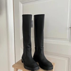 Svarta höga boots från Zara, strl 37 - Säljer ett par svarta höga boots från Zara i storlek 37. De har chunky platt sula med grovt mönster undertill och är tillverkade i syntetmaterial med slät finish. Stövlarna har rund tå och minimalistisk design som ger en cool vibe. Nya med etikett kvar. 