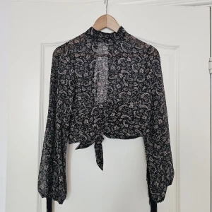 Svart paisley blus med fransar - Säljer en svart blus med paisley-mönster och transparent material. Blusen har långa ärmar med coola fransdetaljer längs sidorna och knyt framtill. Den har en hög hals och croppad passform som ger en edgy vibe. Perfekt för dig som gillar statement-plagg!