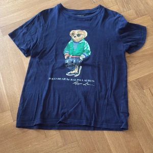 Mörkblå Polo Bear t-shirt Ralph Lauren - Snygg mörkblå t-shirt från Ralph Lauren med Polo Bear-tryck på framsidan. Klassisk rund hals och korta ärmar. Materialet är mjuk bomull och passformen är normal. 