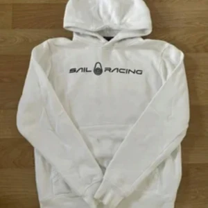 Vit hoodie från Sail Racing - Vit hoodie från Sail Racing med svart logotyptryck på bröstet. Tröjan har huva, lång ärm och en stor magficka framtill. Perfekt för dig som gillar enkel och clean streetstil. Mjuk bomullskänsla och klassisk passform. Storlek 140 på tröjan 