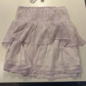 Ljuslila volangkjol med smock - Supersöt kort kjol i ljuslila med bred smock i midjan och dubbla volanger. Kjolen är luftig och har en strechig midja. Dubbelt tyg vilket gör att kjolen INTE är genomskinlig. aldrig använd