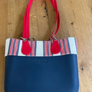 Randig axelväska från O bag - Supersnygg oBag, ca 35cm bred 29cm hög Mörkblå med blå/grå innerväska med ett litet fack. Röda långa axelremmar, röd/vit/blå dekor. Knappt använd