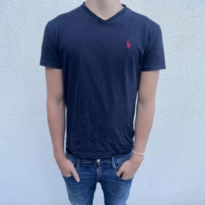 Mörkblå v-ringad t-shirt från Polo Ralph Lauren - Snygg mörkblå t-shirt från Polo Ralph Lauren med klassisk röd logga broderad på bröstet. T-shirten har v-ringad hals och korta ärmar, perfekt för en chill och stilren look. Tillverkad i mjuk bomull som känns skön mot huden.