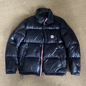 Svart Moncler pufferjacka herr - Svart pufferjacka från Moncler med glansig finish och klassisk logga på bröstet. Jackan har hög krage, dragkedja med röd och vit detalj, samt flera fickor med dragkedja. Perfekt för kalla dagar och har en riktigt schysst streetstil. Jackan är äkta och de står jag för, om private och fråga om vad som helst