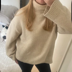 Beige polotröja i stickad ull - Säljer en beige polotröja med bred ribbad krage och muddar. Tröjan är stickad i mjuk ull och har en loose passform med långa ärmar. Perfekt för att hålla sig varm och snygg under kyliga dagar. Enkel och stilren design som är lätt att matcha med jeans eller leggings.