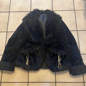 Svart pälsjacka - Säljer en svart pälsjacka med fluffig krage och muddar. Jackan har ett snyggt vågigt mönster i pälsen och coola bältesdetaljer vid midjan. Insidan är quiltad för extra komfort. Perfekt för dig som vill sticka ut med en lyxig vibe.