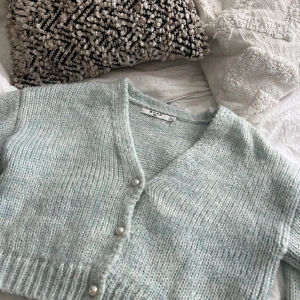 Ljusblå croppad kofta från NA-KD - Supermysig ljusblå stickad kofta från NA-KD med v-ringning och pärlknappar framtill. Croppad modell med långa ärmar och ribbade muddar. Perfekt att slänga på över ett linne eller t-shirt för en chill look.