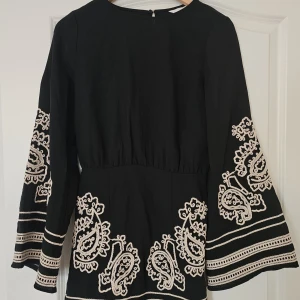 Svart kortklänning med broderi - Svart kortklänning med vita broderade paisleymönster och breda, långa ärmar. Klänningen har markerad midja och dekorativa ränder längst ner på ärmar och kjol. Materialet känns som bomull och ger en skön och avslappnad vibe.