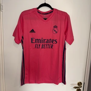 Rosa Real Madrid fotbollströja Adidas M - Snygg rosa Real Madrid fotbollströja från Adidas med mörkblå detaljer och logga. Tröjan har V-ringning, korta ärmar och är gjord i lätt, ventilerande Aeroready-material. Tryck med Emirates Fly Better på bröstet och klassiska Adidas-ränder längs sidorna.
