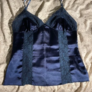 Mörkblå satin linnetopp med spets - Supersnygg mörkblå linnetopp i satin med tunna axelband och v-ringad hals. Toppen har spetsdetaljer längs bysten och sidorna som ger en extra lyxig känsla. Sval och glansig finish, perfekt för dig som gillar en chic och feminin stil.