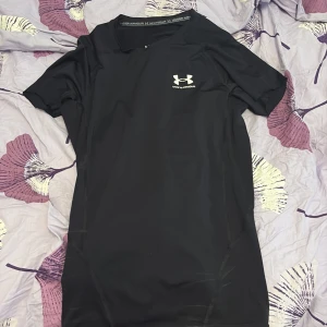 Svart kompressionst-shirt Under Armour - Svart kompressionst-shirt från Under Armour med vit logga på bröstet. T-shirten är kortärmad och har en tight passform, perfekt för träning. Materialet är polyester och designen är stilren utan mönster. Snygg att ha på gymmet eller till sport.