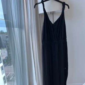 Svart maxiklänning från Lindex - Svart långklänning från Lindex med omlott framtill och twistade axelband. Klänningen har en snygg öppen rygg med knutdetalj och är tillverkad i mjukt material. Perfekt för dig som gillar stilrena och bekväma plagg med coola detaljer.