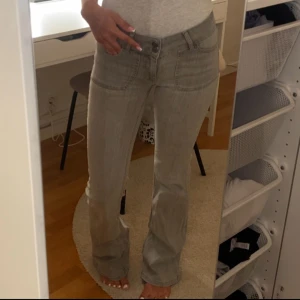 Grå bootcut jeans med fickdetaljer - Snygga grå jeans med bootcut passform och låg midja. Jeansen har breda framfickor och bakfickor med lock, vilket ger en cool och unik look. Tillverkade i klassiskt jeanstyg som är mjukt och följsamt. Perfekta för dig som gillar en avslappnad men trendig stil.