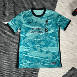 Liverpool FC blå Nike fotbollströja - Snygg Liverpool FC fotbollströja från Nike i en ljusblå färg med mörkblå detaljer. Tröjan har ett unikt abstrakt mönster, svart Nike-logga och LFC-emblem på bröstet. Rund hals med svart kant och svarta muddar på ärmarna. Tillverkad i lätt och ventilerande polyester. Storlek XL barn men passar S vuxen 