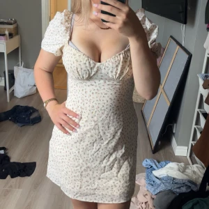 Blommig kortklänning med puffärm - Supersöt kortklänning i ljus beige med små blommor och puffiga kortärmar. Klänningen har en v-ringad urringning och sitter tight över midjan för en feminin siluett. Perfekt för sommarens alla tillfällen, lätt och skön i bomull.