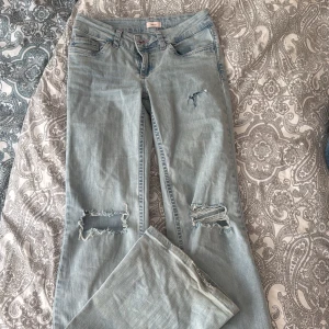 Bootcut ljusblå jeans från Nelly - Säljer ett par ljusblå bootcut jeans från Nelly med slitna detaljer och hål vid knäna. Hålet på höger sida högre upp var ett misstag men tycker ändå det ser snyggt ut! Ganska slitna längst ner vid benen men annars inget fel. Säljer för att dom inte kommer till användning❣️storlek 36!