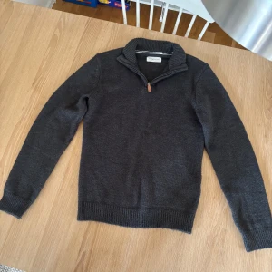 Mörkgrå stickad tröja Pier One - Mörkgrå stickad tröja från Pier One med ribbad krage och half zip-dragkedja. Tröjan har långärmade ärmar och är gjord i mjuk blandning av bomull och syntet. Perfekt för kyliga dagar och enkel att matcha med jeans eller chinos.